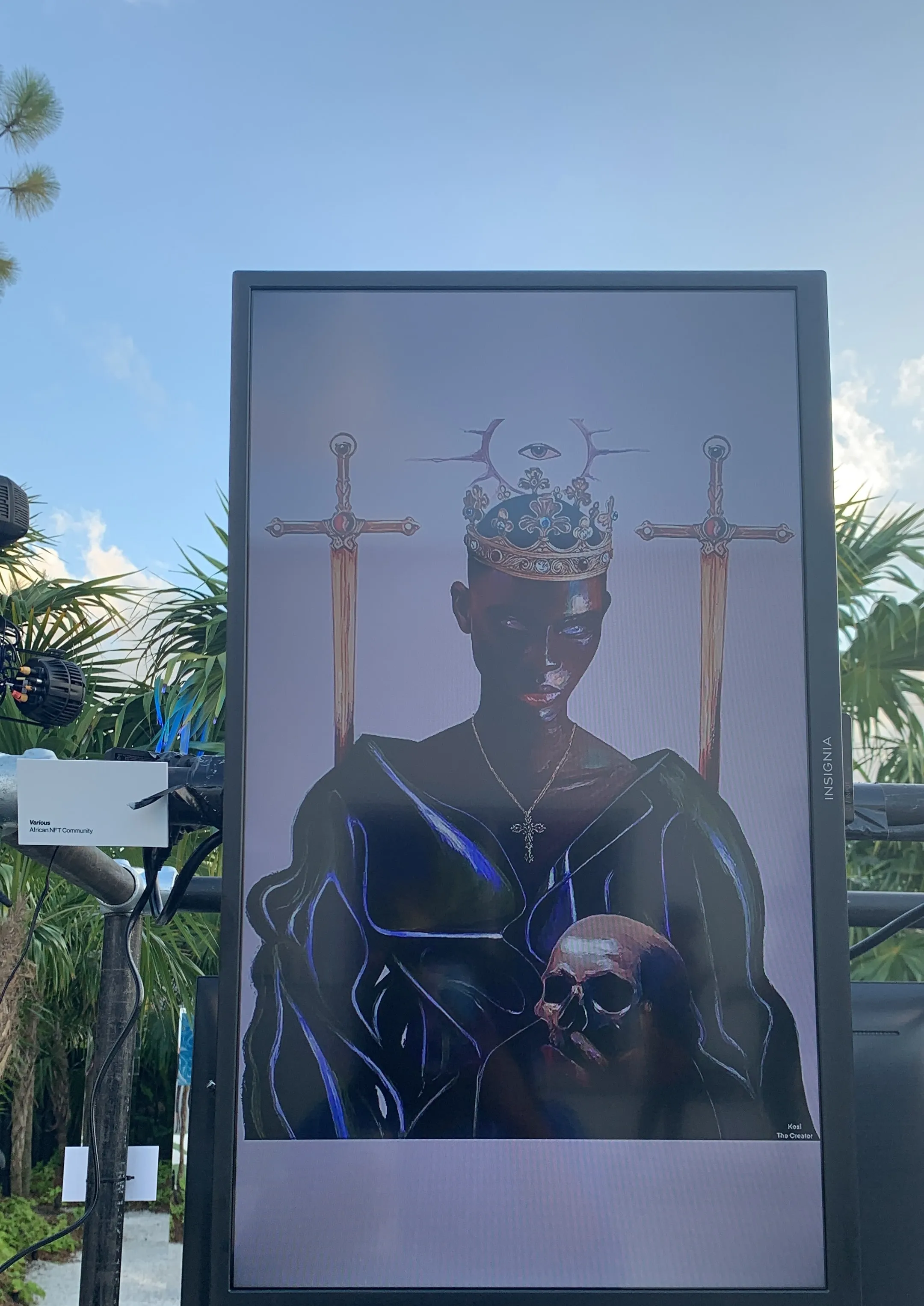 King's Secret on a screen turned portrait, mounted on a stand outdoors in daylight, with palms and blue sky behind it. A small printed label to the left reads Various, African NFT Community.