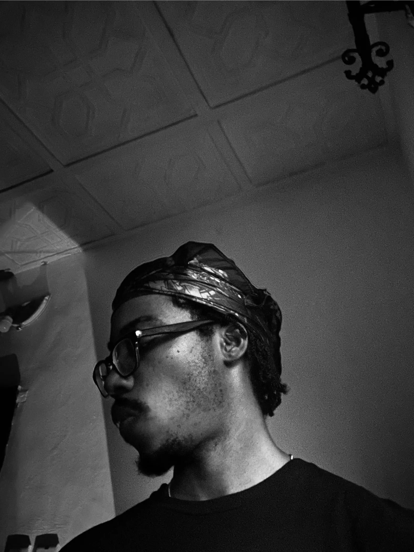 Kosi Okafor in a patterned head wrap and wire-rimmed glasses, head turned away from the camera. Black and white, shot from below in low light.